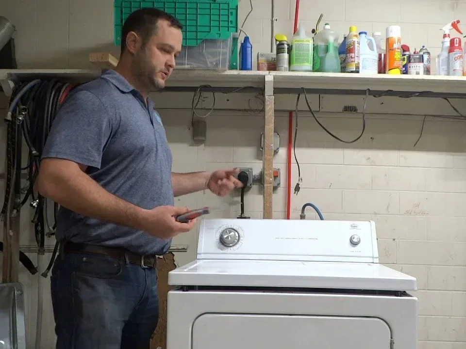 Technician checking the power cord and outlet behind a white electric dryer in a workshop